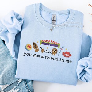 May include: Light blue sweatshirt with embroidered Toy Story-themed graphics. Features a ball, boot, roller coaster, dog, and cowboy hat. The text "You got a friend in me" is embroidered below the graphics. A silver necklace is draped across the sweatshirt.