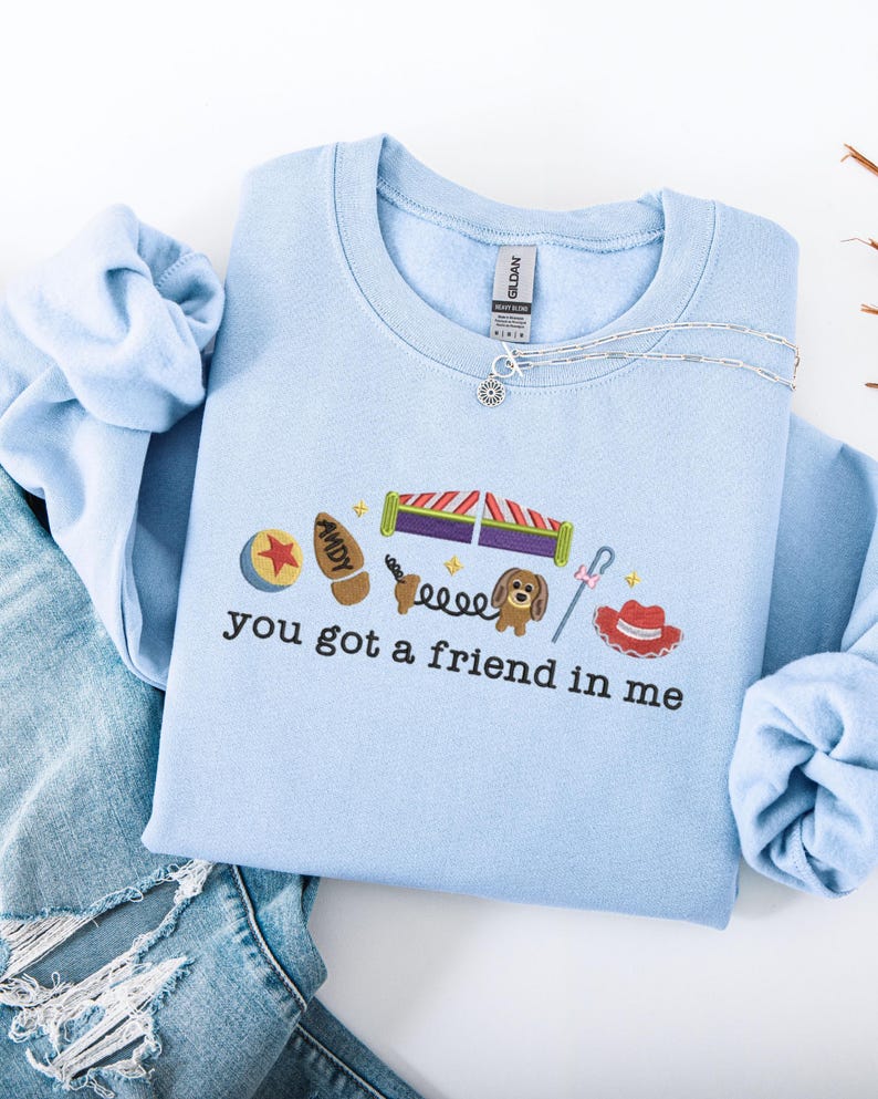 May include: Light blue sweatshirt with embroidered Toy Story-themed graphics. Features a ball, boot, roller coaster, dog, and cowboy hat. The text "You got a friend in me" is embroidered below the graphics. A silver necklace is draped across the sweatshirt.