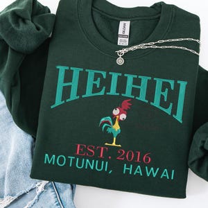 May include: Dark green sweatshirt with the word "HEIHEI" in teal lettering. Below the word is a cartoon rooster and the text "EST. 2016 MOTUNUI, HAWAII" in red and teal. A silver necklace with a sun pendant is on the sweatshirt.