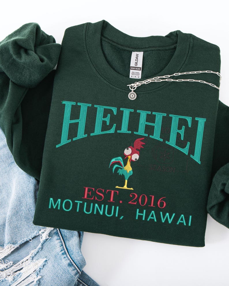 May include: Dark green sweatshirt with the word "HEIHEI" in teal lettering. Below the word is a cartoon rooster and the text "EST. 2016 MOTUNUI, HAWAII" in red and teal. A silver necklace with a sun pendant is on the sweatshirt.
