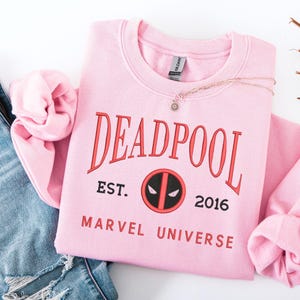 May include: A pale pink crew neck sweatshirt featuring the word "DEADPOOL" in red. Below the word are the words "EST. 2016" and a circular logo. The words "MARVEL UNIVERSE" are printed below the logo. A gold necklace with a small pendant is on the sweatshirt.