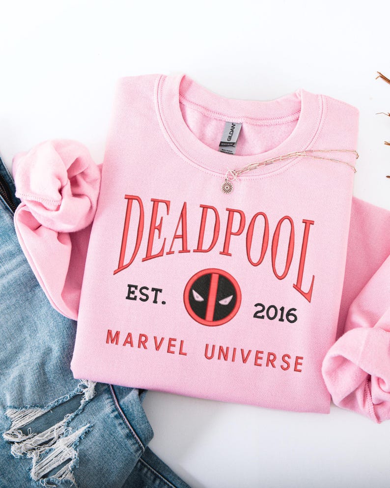 May include: A pale pink crew neck sweatshirt featuring the word "DEADPOOL" in red. Below the word are the words "EST. 2016" and a circular logo. The words "MARVEL UNIVERSE" are printed below the logo. A gold necklace with a small pendant is on the sweatshirt.