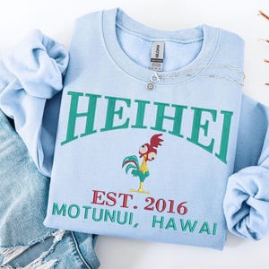 May include: Light blue sweatshirt with the word "HEIHEI" in green arched lettering. Below the text is a cartoon rooster and the text "EST. 2016 MOTUNUI, HAWAII" in red and green. A silver necklace is draped across the sweatshirt.