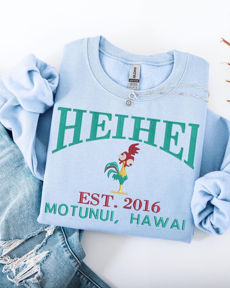 May include: Light blue sweatshirt with the word "HEIHEI" in green arched lettering. Below the text is a cartoon rooster and the text "EST. 2016 MOTUNUI, HAWAII" in red and green. A silver necklace is draped across the sweatshirt.