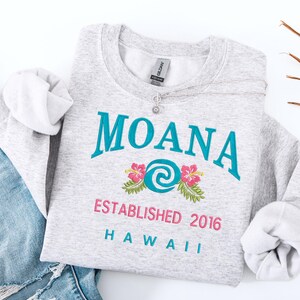 K&ouml;nnte beinhalten: Ein meliertes graues Sweatshirt mit dem Wort "MOANA" in T&uuml;rkis, einem Blumendesign und dem Text "ESTABLISHED 2016 HAWAII" in Pink und T&uuml;rkis. Eine silberne Halskette mit Blumenanh&auml;nger liegt darauf.