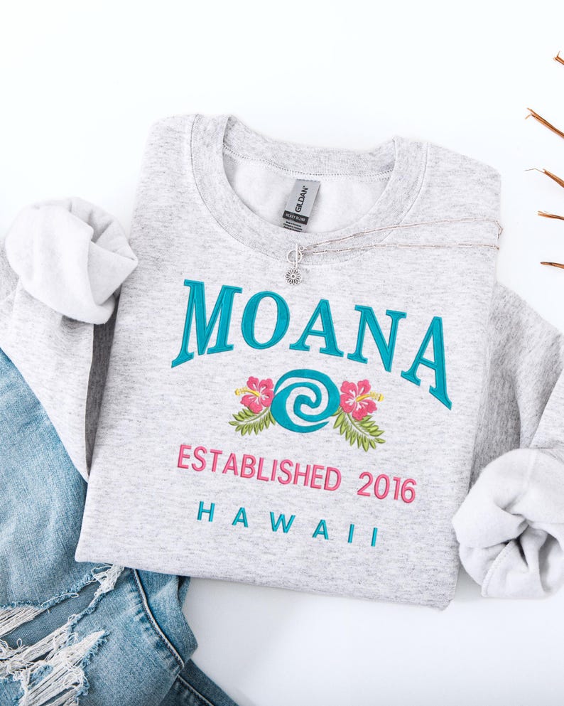 K&ouml;nnte beinhalten: Ein meliertes graues Sweatshirt mit dem Wort "MOANA" in T&uuml;rkis, einem Blumendesign und dem Text "ESTABLISHED 2016 HAWAII" in Pink und T&uuml;rkis. Eine silberne Halskette mit Blumenanh&auml;nger liegt darauf.