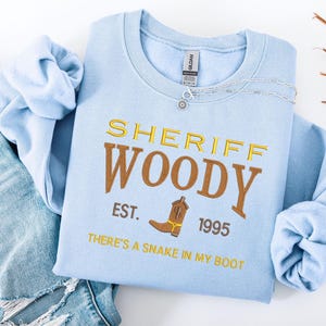 Puede incluir: Sudadera azul claro con la inscripci&oacute;n "SHERIFF WOODY" bordada en dorado. Debajo, se lee "EST. 1995" con un dibujo de bota de vaquero marr&oacute;n. La frase "THERE'S A SNAKE IN MY BOOT" tambi&eacute;n est&aacute; bordada en dorado.