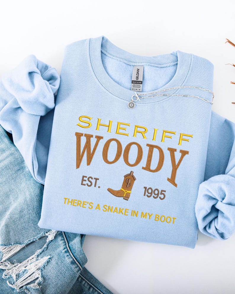 Puede incluir: Sudadera azul claro con la inscripci&oacute;n "SHERIFF WOODY" bordada en dorado. Debajo, se lee "EST. 1995" con un dibujo de bota de vaquero marr&oacute;n. La frase "THERE'S A SNAKE IN MY BOOT" tambi&eacute;n est&aacute; bordada en dorado.