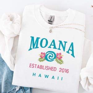 K&ouml;nnte beinhalten: Wei&szlig;es Sweatshirt mit Rundhalsausschnitt, auf dem das Wort "MOANA" in T&uuml;rkis, ein Blumendesign und der Text "ESTABLISHED 2016 HAWAII" in Rosa und T&uuml;rkis zu sehen sind. Eine silberne Halskette mit einem Blumenanh&auml;nger liegt auf dem Sweatshirt.