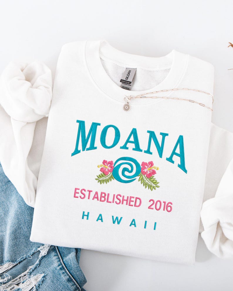 K&ouml;nnte beinhalten: Wei&szlig;es Sweatshirt mit Rundhalsausschnitt, auf dem das Wort "MOANA" in T&uuml;rkis, ein Blumendesign und der Text "ESTABLISHED 2016 HAWAII" in Rosa und T&uuml;rkis zu sehen sind. Eine silberne Halskette mit einem Blumenanh&auml;nger liegt auf dem Sweatshirt.