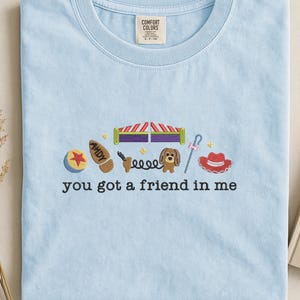May include: Light blue t-shirt with embroidered images of a ball, boot, circus tent, dog, and cowboy hat. The text "you got a friend in me" is embroidered below the images. The shirt has a crew neck and a tag that says "Comfort Colors".