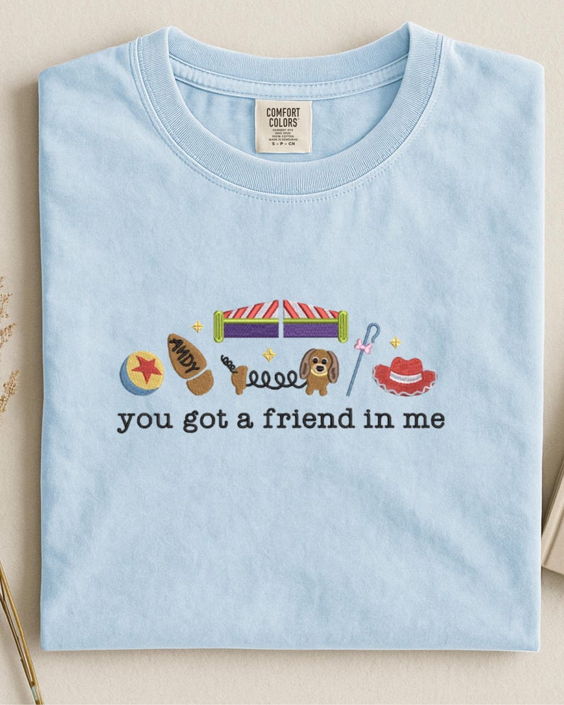 May include: Light blue t-shirt with embroidered images of a ball, boot, circus tent, dog, and cowboy hat. The text "you got a friend in me" is embroidered below the images. The shirt has a crew neck and a tag that says "Comfort Colors".