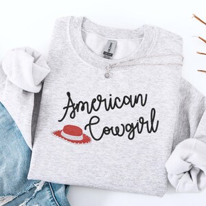 May include: A light grey sweatshirt featuring the text "American Cowgirl" in black script, with a red and white cowboy hat graphic. A silver necklace with a flower pendant is visible. The sweatshirt is paired with blue denim jeans.