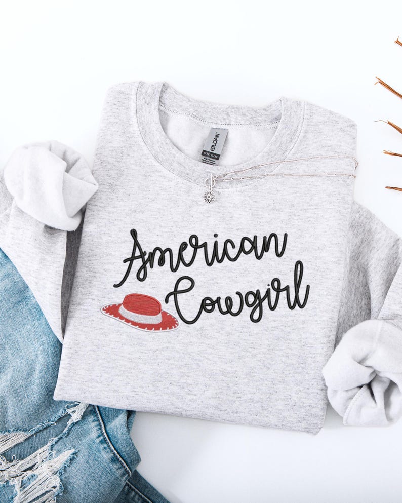 May include: A light grey sweatshirt featuring the text "American Cowgirl" in black script, with a red and white cowboy hat graphic. A silver necklace with a flower pendant is visible. The sweatshirt is paired with blue denim jeans.