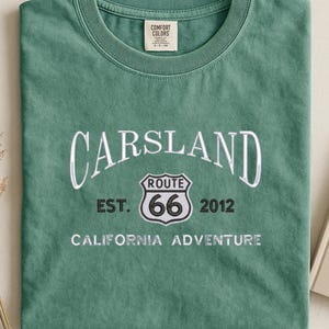 Pode incluir: Uma t-shirt verde com a palavra "CARSLAND" em escrita branca. Abaixo, um logótipo da Rota 66 com o número "66" no interior. O texto "EST. 2012 CALIFORNIA ADVENTURE" está por baixo. Uma etiqueta Comfort Colors é visível.