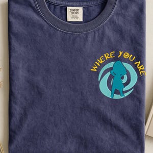 Może przedstawiać: Granatowa koszulka z napisem "WHERE YOU ARE" w kolorze ż&oacute;łtym i turkusową grafiką. Koszulka ma okrągły dekolt i małą metkę na kołnierzu.