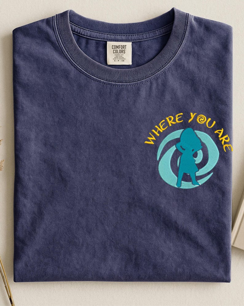 Może przedstawiać: Granatowa koszulka z napisem "WHERE YOU ARE" w kolorze ż&oacute;łtym i turkusową grafiką. Koszulka ma okrągły dekolt i małą metkę na kołnierzu.