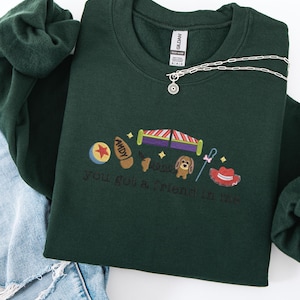 May include: Dark green sweatshirt with embroidered Toy Story characters and the phrase "you got a friend in me." The design includes a ball, a boot, a rocket, a dog, a cane, and a cowboy hat. A silver necklace with a sun pendant is draped on the sweatshirt.