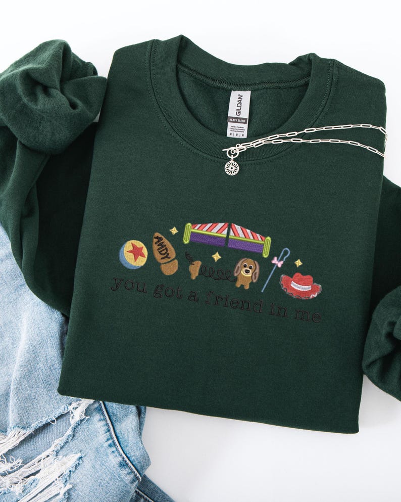 May include: Dark green sweatshirt with embroidered Toy Story characters and the phrase "you got a friend in me." The design includes a ball, a boot, a rocket, a dog, a cane, and a cowboy hat. A silver necklace with a sun pendant is draped on the sweatshirt.
