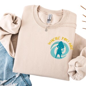 Może przedstawiać: Beżowa bluza z napisem "WHERE YOU ARE" i turkusową grafiką sylwetki w wirującym wzorze. Srebrny naszyjnik z zawieszką w kształcie słońca leży na bluzie. Bluza jest złożona na parze niebieskich dżins&oacute;w.