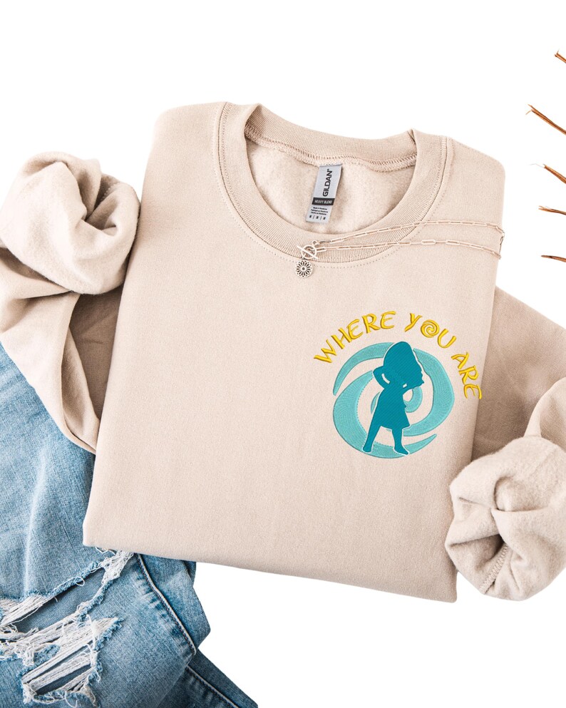 Może przedstawiać: Beżowa bluza z napisem "WHERE YOU ARE" i turkusową grafiką sylwetki w wirującym wzorze. Srebrny naszyjnik z zawieszką w kształcie słońca leży na bluzie. Bluza jest złożona na parze niebieskich dżins&oacute;w.