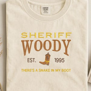 Puede incluir: Camiseta crema con las palabras "SHERIFF WOODY" en letras doradas y marrones, un dibujo de bota de vaquero marr&oacute;n y los textos "EST. 1995" y "THERE'S A SNAKE IN MY BOOT". La camiseta est&aacute; doblada.