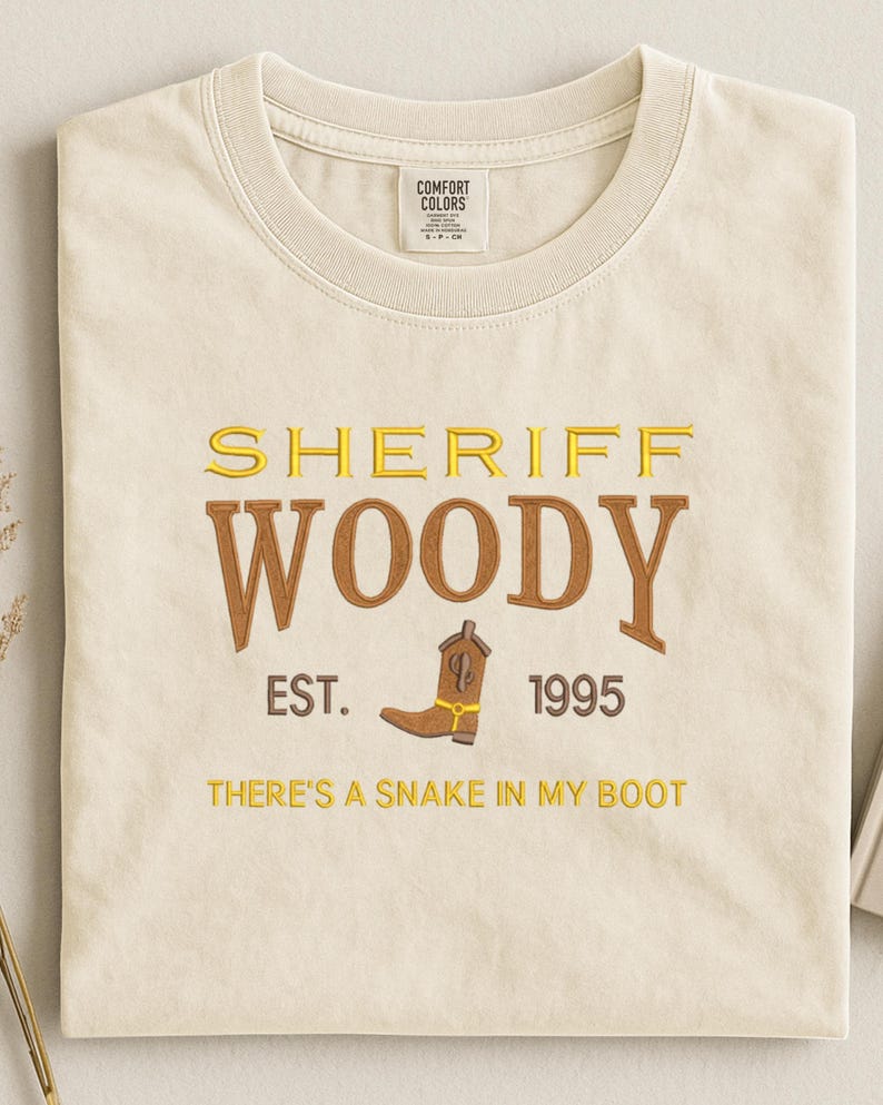 Puede incluir: Camiseta crema con las palabras "SHERIFF WOODY" en letras doradas y marrones, un dibujo de bota de vaquero marr&oacute;n y los textos "EST. 1995" y "THERE'S A SNAKE IN MY BOOT". La camiseta est&aacute; doblada.
