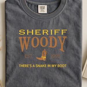 Puede incluir: Una camiseta gris carb&oacute;n con las palabras "SHERIFF WOODY" en letras amarillas y marrones. Debajo del texto hay un gr&aacute;fico de bota de vaquero marr&oacute;n y la frase "THERE'S A SNAKE IN MY BOOT". La camiseta tambi&eacute;n tiene el texto "EST. 1995".