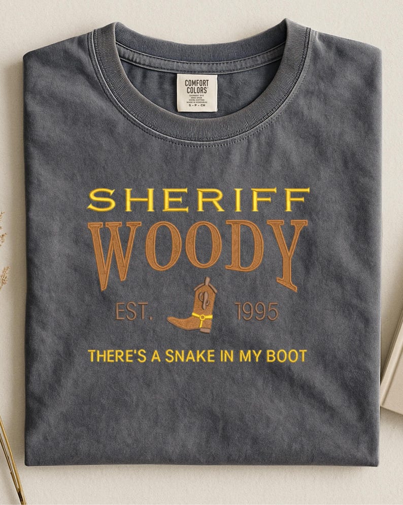 Puede incluir: Una camiseta gris carb&oacute;n con las palabras "SHERIFF WOODY" en letras amarillas y marrones. Debajo del texto hay un gr&aacute;fico de bota de vaquero marr&oacute;n y la frase "THERE'S A SNAKE IN MY BOOT". La camiseta tambi&eacute;n tiene el texto "EST. 1995".