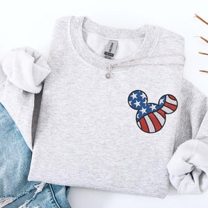 Peut inclure: Un sweat-shirt gris chin&eacute; avec un motif Mickey Mouse patriotique. Le motif reprend les couleurs du drapeau am&eacute;ricain : rayures rouges et blanches et &eacute;toiles bleues. Un collier argent&eacute; avec un pendentif en forme de fleur est &eacute;galement visible.