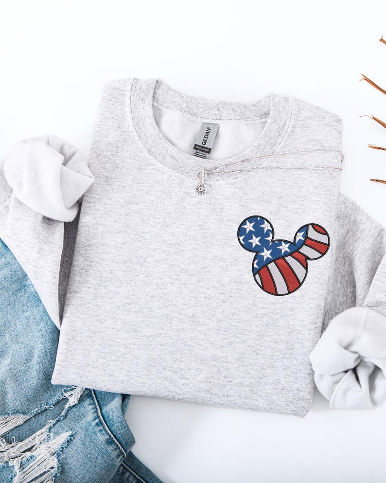 Peut inclure: Un sweat-shirt gris chin&eacute; avec un motif Mickey Mouse patriotique. Le motif reprend les couleurs du drapeau am&eacute;ricain : rayures rouges et blanches et &eacute;toiles bleues. Un collier argent&eacute; avec un pendentif en forme de fleur est &eacute;galement visible.