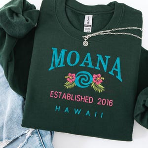K&ouml;nnte beinhalten: Dunkelgr&uuml;nes Sweatshirt mit dem Wort "MOANA" in T&uuml;rkis, rosa Blumen und dem Text "ESTABLISHED 2016 HAWAII". Eine silberne Halskette mit Sonnenanh&auml;nger liegt auf dem Sweatshirt. Das Sweatshirt ist auf einer Jeans gefaltet.