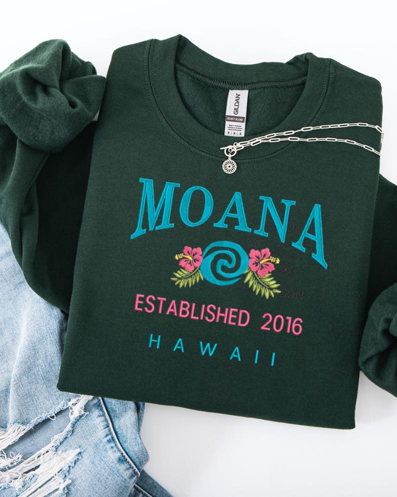 K&ouml;nnte beinhalten: Dunkelgr&uuml;nes Sweatshirt mit dem Wort "MOANA" in T&uuml;rkis, rosa Blumen und dem Text "ESTABLISHED 2016 HAWAII". Eine silberne Halskette mit Sonnenanh&auml;nger liegt auf dem Sweatshirt. Das Sweatshirt ist auf einer Jeans gefaltet.