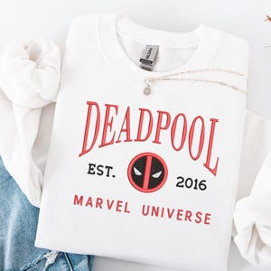 May include: White crew neck sweatshirt featuring the word "DEADPOOL" in red, with "EST. 2016" and "MARVEL UNIVERSE" printed below. A silver necklace with a small pendant is placed on the sweatshirt. The sweatshirt is folded on blue denim.
