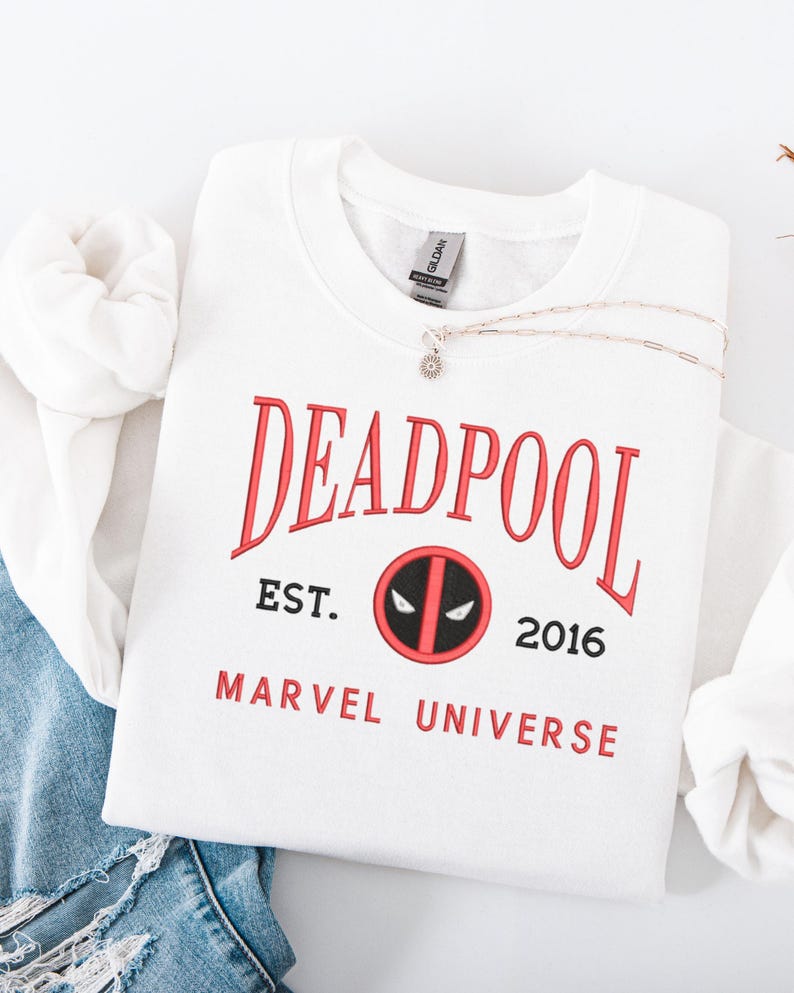 May include: White crew neck sweatshirt featuring the word "DEADPOOL" in red, with "EST. 2016" and "MARVEL UNIVERSE" printed below. A silver necklace with a small pendant is placed on the sweatshirt. The sweatshirt is folded on blue denim.