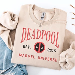 May include: A beige crew neck sweatshirt featuring the word "DEADPOOL" in red lettering. Below the word is "EST. 2016" and the Marvel Universe logo. A silver necklace with a sun pendant is on the sweatshirt.