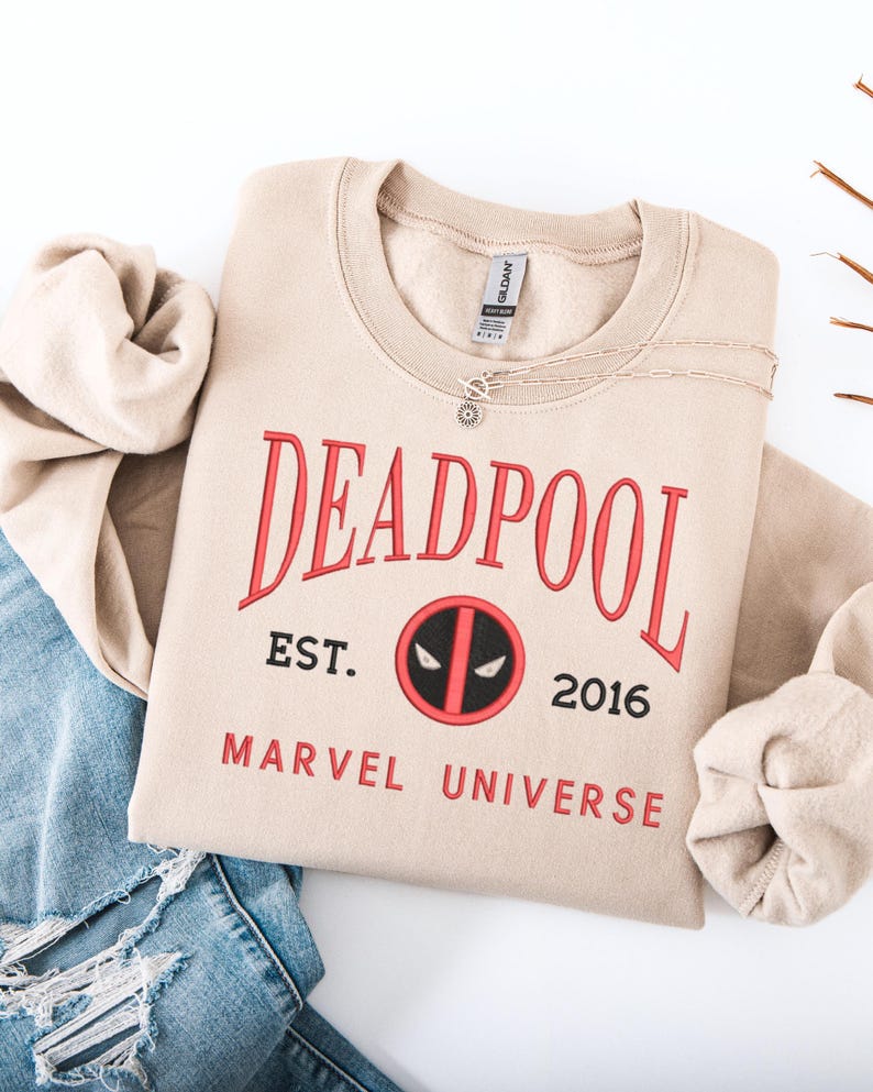May include: A beige crew neck sweatshirt featuring the word "DEADPOOL" in red lettering. Below the word is "EST. 2016" and the Marvel Universe logo. A silver necklace with a sun pendant is on the sweatshirt.