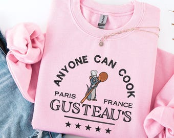 May include: A light pink sweatshirt with the text "ANYONE CAN COOK" in a semi-circle above "GUSTEAU'S PARIS FRANCE" and a cartoon rat chef holding a wooden spoon. The sweatshirt is paired with blue jeans.