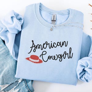 May include: A pale blue sweatshirt featuring the text "American Cowgirl" in black script, and a red and white cowboy hat graphic. A silver necklace is draped across the front. The sweatshirt is folded on top of a pair of blue jeans.