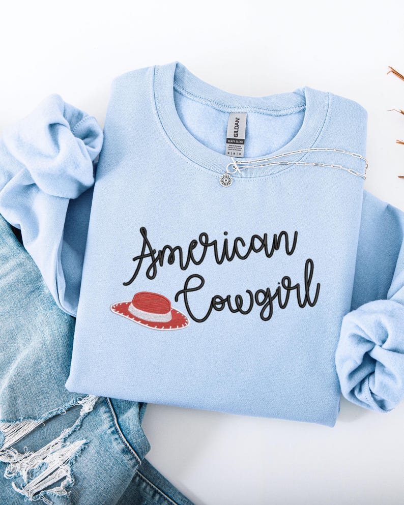 May include: A pale blue sweatshirt featuring the text "American Cowgirl" in black script, and a red and white cowboy hat graphic. A silver necklace is draped across the front. The sweatshirt is folded on top of a pair of blue jeans.