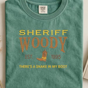 Puede incluir: Camiseta verde con el texto "SHERIFF WOODY" en amarillo y marr&oacute;n, junto con un gr&aacute;fico de bota de vaquero. La camiseta tambi&eacute;n incluye el texto "EST. 1995" y "THERE'S A SNAKE IN MY BOOT".