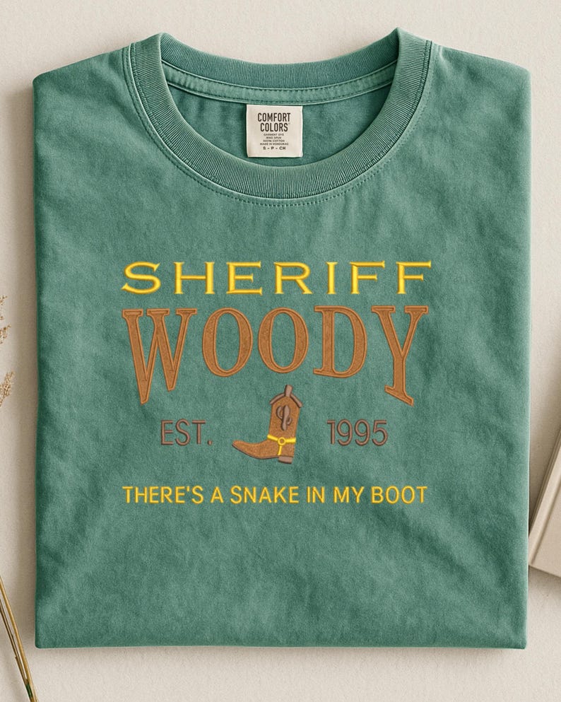 Puede incluir: Camiseta verde con el texto "SHERIFF WOODY" en amarillo y marr&oacute;n, junto con un gr&aacute;fico de bota de vaquero. La camiseta tambi&eacute;n incluye el texto "EST. 1995" y "THERE'S A SNAKE IN MY BOOT".