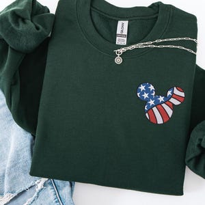 Peut inclure: Un sweat-shirt vert fonc&eacute; avec un motif Mickey Mouse aux couleurs du drapeau am&eacute;ricain. Le motif pr&eacute;sente des rayures rouges et blanches et des &eacute;toiles bleues. Un collier argent&eacute; avec un pendentif soleil est visible.