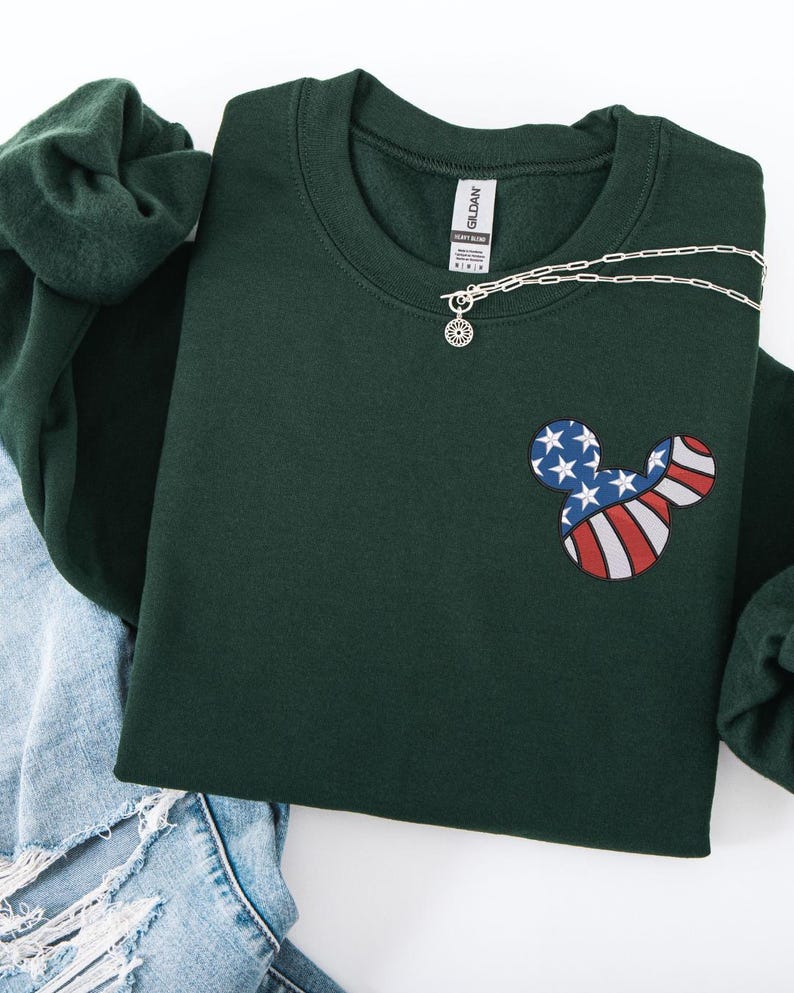 Peut inclure: Un sweat-shirt vert fonc&eacute; avec un motif Mickey Mouse aux couleurs du drapeau am&eacute;ricain. Le motif pr&eacute;sente des rayures rouges et blanches et des &eacute;toiles bleues. Un collier argent&eacute; avec un pendentif soleil est visible.