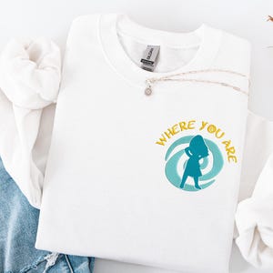 Może przedstawiać: Biała bluza z okrągłym dekoltem z napisem "WHERE YOU ARE" w kolorze złotym i turkusową grafiką kobiety przed turkusowym wirkiem. Srebrny naszyjnik z wisiorkiem w kształcie kwiatu znajduje się na bluzie. Bluza jest złożona na niebieskim dżinsie.