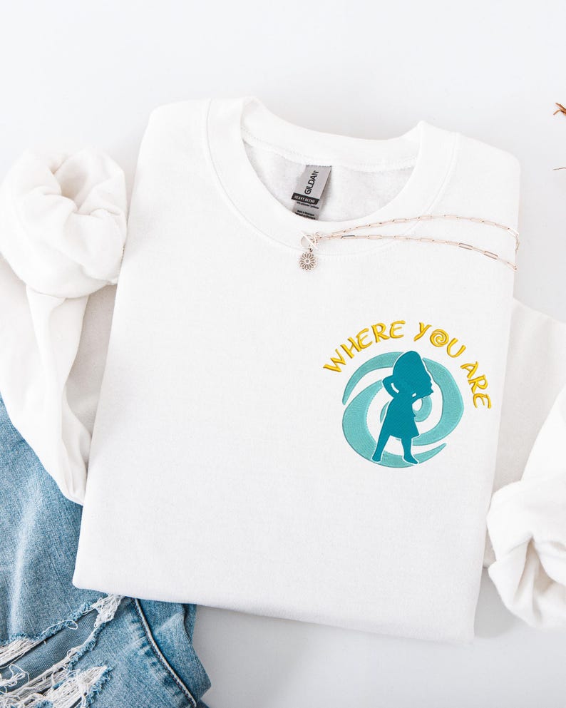 Może przedstawiać: Biała bluza z okrągłym dekoltem z napisem "WHERE YOU ARE" w kolorze złotym i turkusową grafiką kobiety przed turkusowym wirkiem. Srebrny naszyjnik z wisiorkiem w kształcie kwiatu znajduje się na bluzie. Bluza jest złożona na niebieskim dżinsie.
