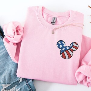 Peut inclure: Un sweat-shirt rose clair &agrave; col rond et manches longues. Le sweat-shirt pr&eacute;sente un motif de t&ecirc;te de Mickey Mouse sur la poitrine, avec un motif de drapeau am&eacute;ricain rouge, blanc et bleu. Un collier en or avec un petit pendentif est drap&eacute; sur le sweat-shirt.