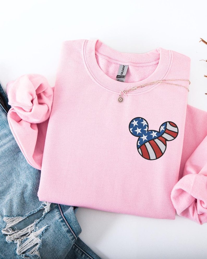 Peut inclure: Un sweat-shirt rose clair &agrave; col rond et manches longues. Le sweat-shirt pr&eacute;sente un motif de t&ecirc;te de Mickey Mouse sur la poitrine, avec un motif de drapeau am&eacute;ricain rouge, blanc et bleu. Un collier en or avec un petit pendentif est drap&eacute; sur le sweat-shirt.