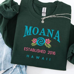 K&ouml;nnte beinhalten: Dunkelgr&uuml;nes Sweatshirt mit dem Wort "MOANA" in T&uuml;rkis, einem t&uuml;rkisfarbenen Wirbeldesign und rosa Hibiskusbl&uuml;ten. Der Text "ESTABLISHED 2016 HAWAII" steht darunter. Eine silberne Halskette und eine dunkelgr&uuml;ne Haargummi sind ebenfalls zu sehen.