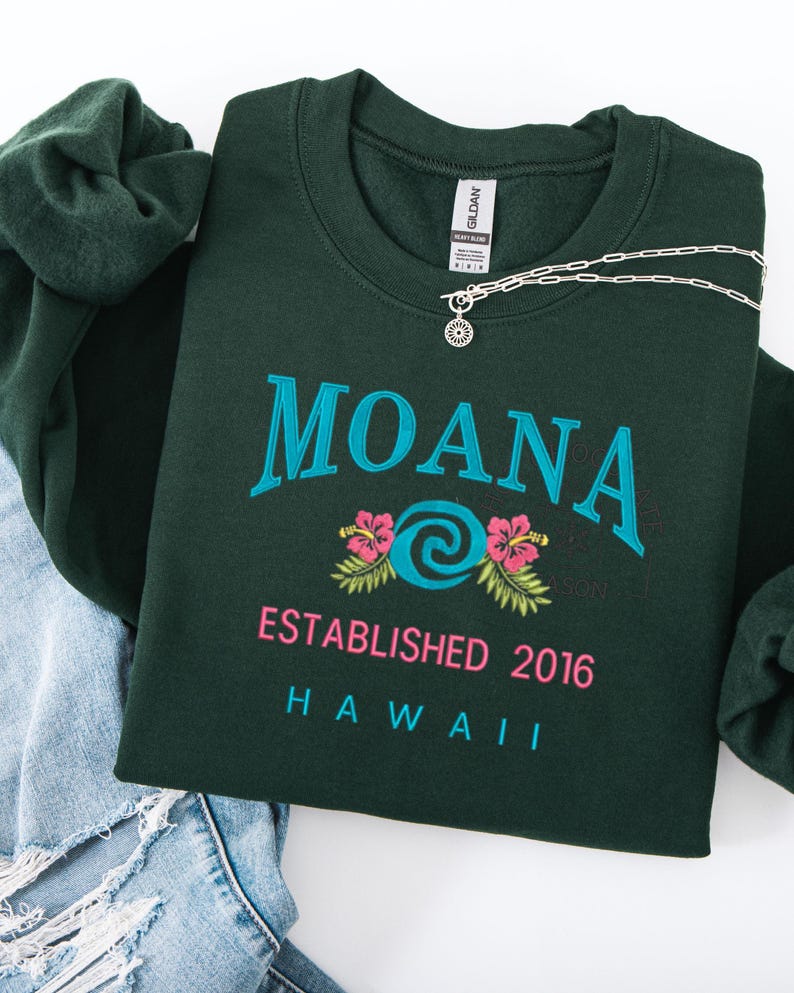 K&ouml;nnte beinhalten: Dunkelgr&uuml;nes Sweatshirt mit dem Wort "MOANA" in T&uuml;rkis, einem t&uuml;rkisfarbenen Wirbeldesign und rosa Hibiskusbl&uuml;ten. Der Text "ESTABLISHED 2016 HAWAII" steht darunter. Eine silberne Halskette und eine dunkelgr&uuml;ne Haargummi sind ebenfalls zu sehen.